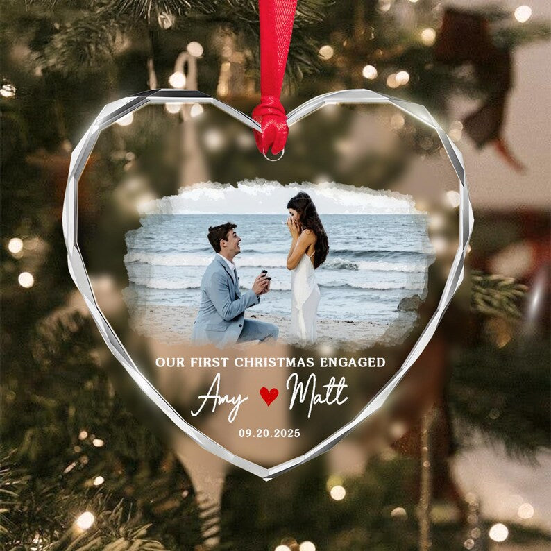 A heart-shaped clear acrylic ornament with faceted edges, featuring a printed photograph of a proposal on a beach. The text reads OUR FIRST CHRISTMAS ENGAGED, Amy, Matt, and 09.20.2025, with a red heart icon.