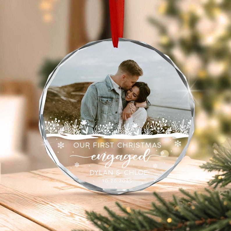 A circular glass ornament with a faceted edge, featuring a photograph of a couple embracing outdoors. The ornament includes white printed text that reads OUR FIRST CHRISTMAS engaged DYLAN & CHLOE 10.26.2025 with snowflake accents.
