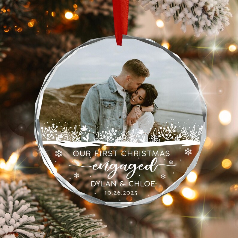 A circular glass Christmas ornament featuring a photograph of a couple embracing outdoors. The ornament includes white printed text that reads "OUR FIRST CHRISTMAS engaged DYLAN & CHLOE 10.26.2025" with a decorative floral border.