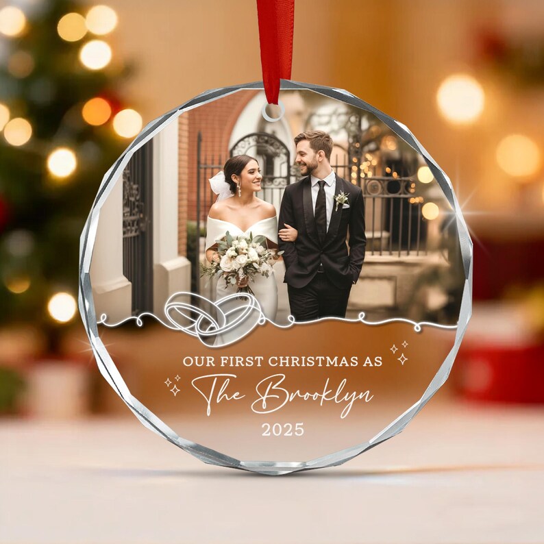 A circular glass Christmas ornament featuring a photo of a couple in wedding attire. The ornament includes a white line drawing of two wedding rings and the text: OUR FIRST CHRISTMAS AS The Brooklyn 2025.