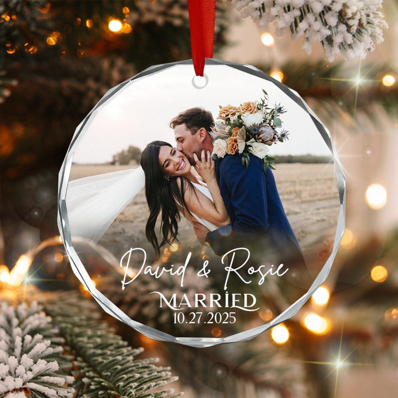 A circular, faceted glass ornament featuring a printed photograph of a couple embracing in a field. The text "David & Rosie MARRIED 10.27.2025" is printed in white script and serif fonts.