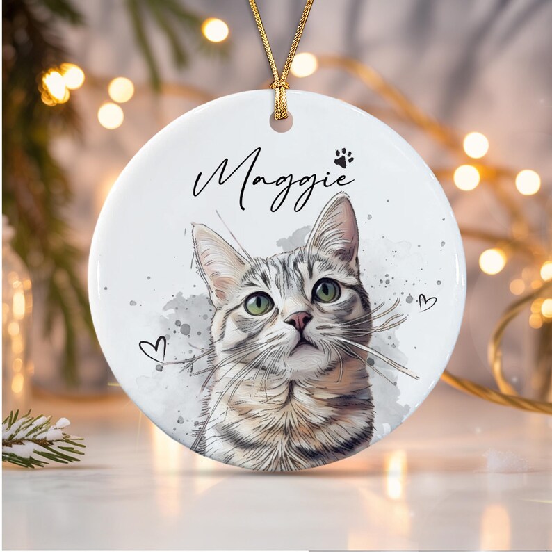 A round white ceramic ornament featuring a watercolor-style illustration of a grey tabby cat with green eyes. The name Maggie is printed in black script above the cat, accompanied by a small paw print and two heart outlines.