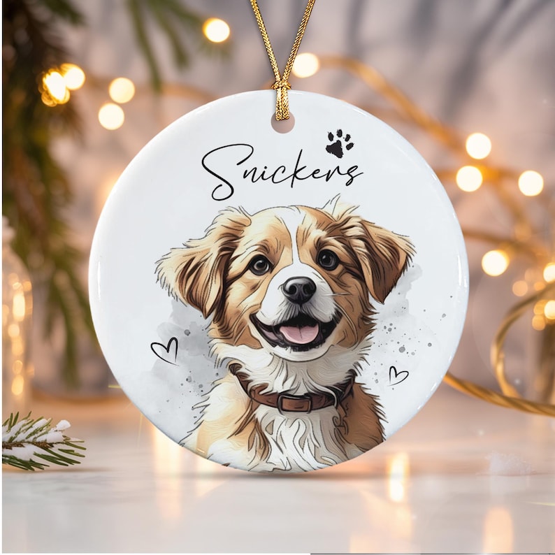 A round white ceramic ornament featuring a watercolor-style illustration of a brown and white dog with a collar. The name Snickers is printed in black script above the dog, accompanied by a small black paw print and two hearts.