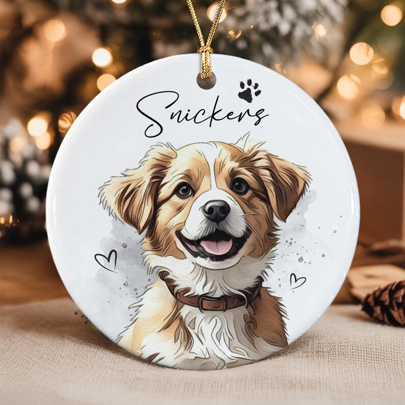 A round white ceramic ornament featuring a colorful illustration of a brown and white dog with a brown collar. The name Snickers is printed in black script above the dog, accompanied by a small paw print and two heart outlines.