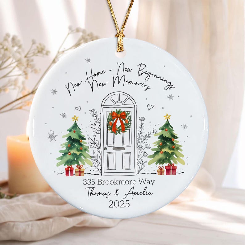 A round white ceramic ornament with a gold string, featuring a line drawing of a front door with a wreath, two Christmas trees with gifts, and the text: New Home - New Beginnings New Memories, 335 Brookmore Way, Thomas & Amelia.
