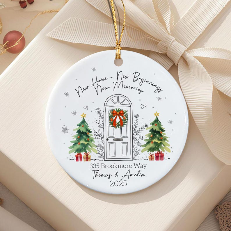 A round white ceramic ornament featuring a central illustration of a front door with a wreath, flanked by two decorated Christmas trees. Text reads: New Home - New Beginnings New Memories, 335 Brookmore Way, Thomas & Amelia, 2025.