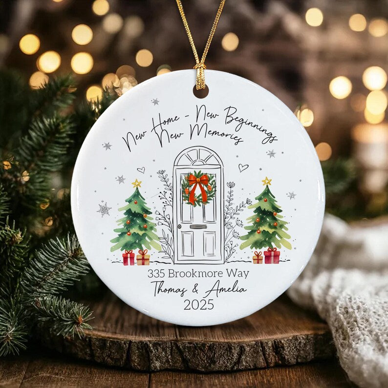 A round white ceramic ornament with a gold string featuring a line drawing of a front door with a wreath, two Christmas trees, and the text New Home - New Beginnings New Memories, 335 Brookmore Way, Thomas & Amelia, and 2025.