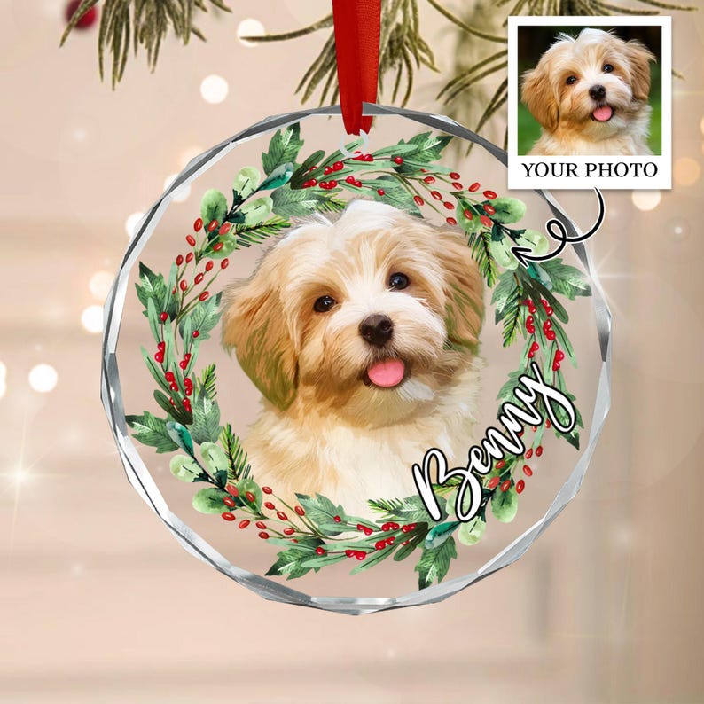 A round, clear acrylic ornament featuring a portrait of a light brown and white puppy centered within a green pine wreath with red berries. The name Benny is written in black script, and a red ribbon is attached to the top.