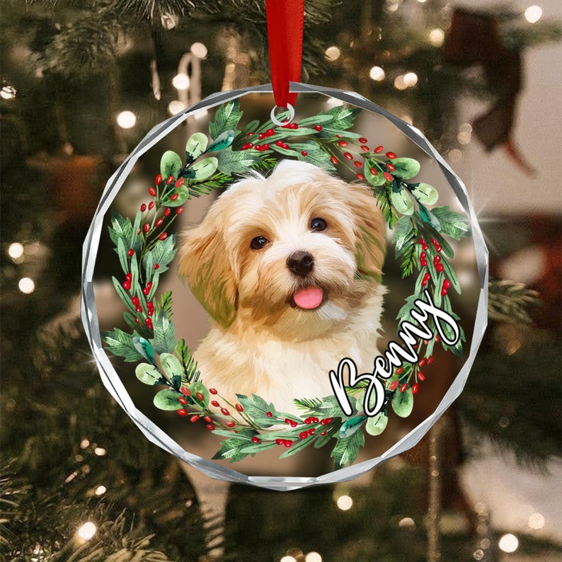 A round, clear acrylic Christmas ornament featuring a printed illustration of a fluffy, light-brown and white puppy centered within a green wreath with red berries.