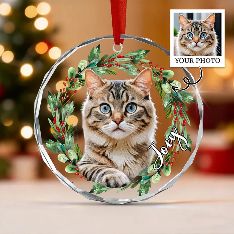 A circular clear acrylic ornament featuring a printed portrait of a brown tabby cat with blue eyes, surrounded by a watercolor-style green holly wreath with red berries.