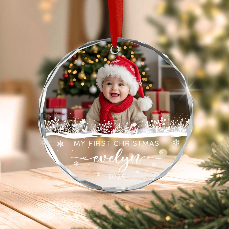 A circular glass Christmas ornament featuring a photo of a baby wearing a red Santa hat and scarf. The ornament includes white printed text that reads MY FIRST CHRISTMAS, the name evelyn, and the year 2025, with snowflake accents.