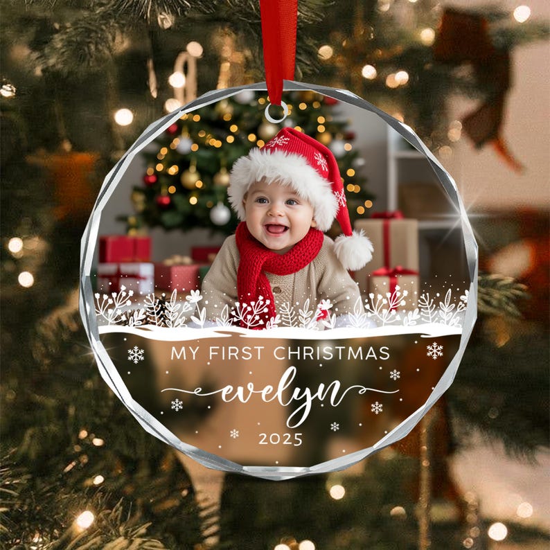 A round, faceted glass Christmas ornament featuring a photo of a baby wearing a red Santa hat and scarf.