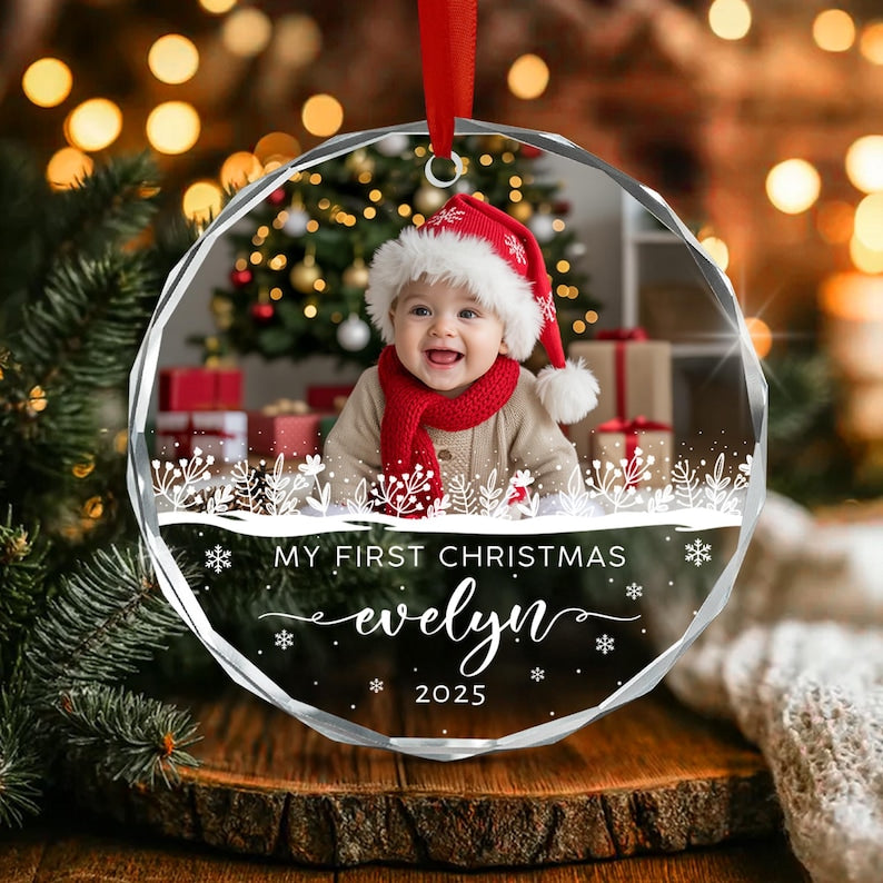 A round, clear acrylic Christmas ornament featuring a photograph of a baby wearing a red and white Santa hat and scarf. The ornament includes white printed text that reads MY FIRST CHRISTMAS, evelyn, and the year 2025.