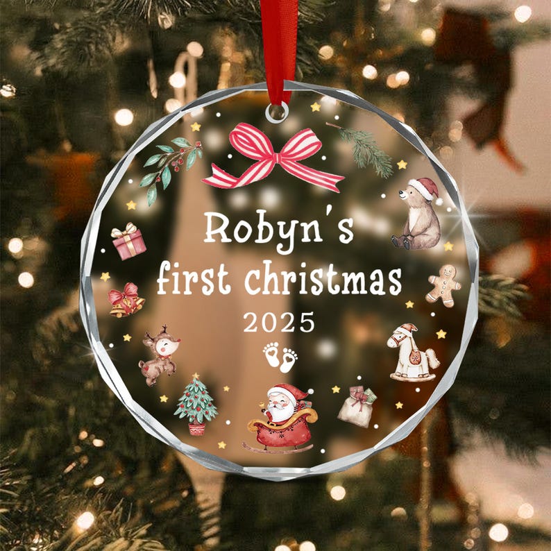 A round, clear, faceted glass ornament hanging from a red ribbon. It features a red and white striped bow at the top and the text 'Robyn's first christmas 2025'. Illustrations include a bear, gingerbread man, Santa, and reindeer.
