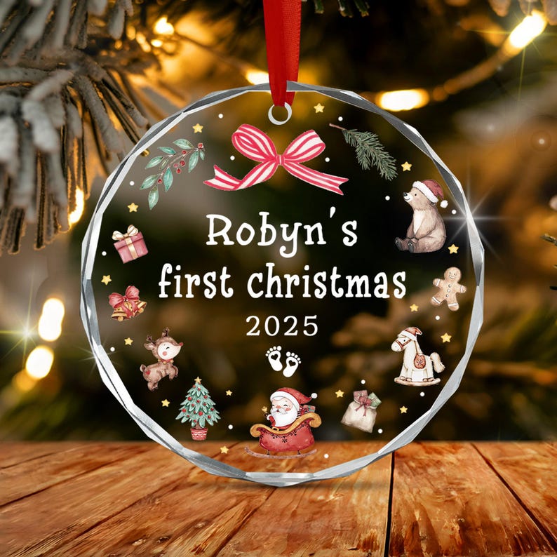 A round, faceted clear acrylic Christmas ornament hanging from a red ribbon. It features the text "Robyn's first christmas 2025" with illustrations of a bear, gingerbread man, rocking horse, Santa in a sleigh, and baby footprints.