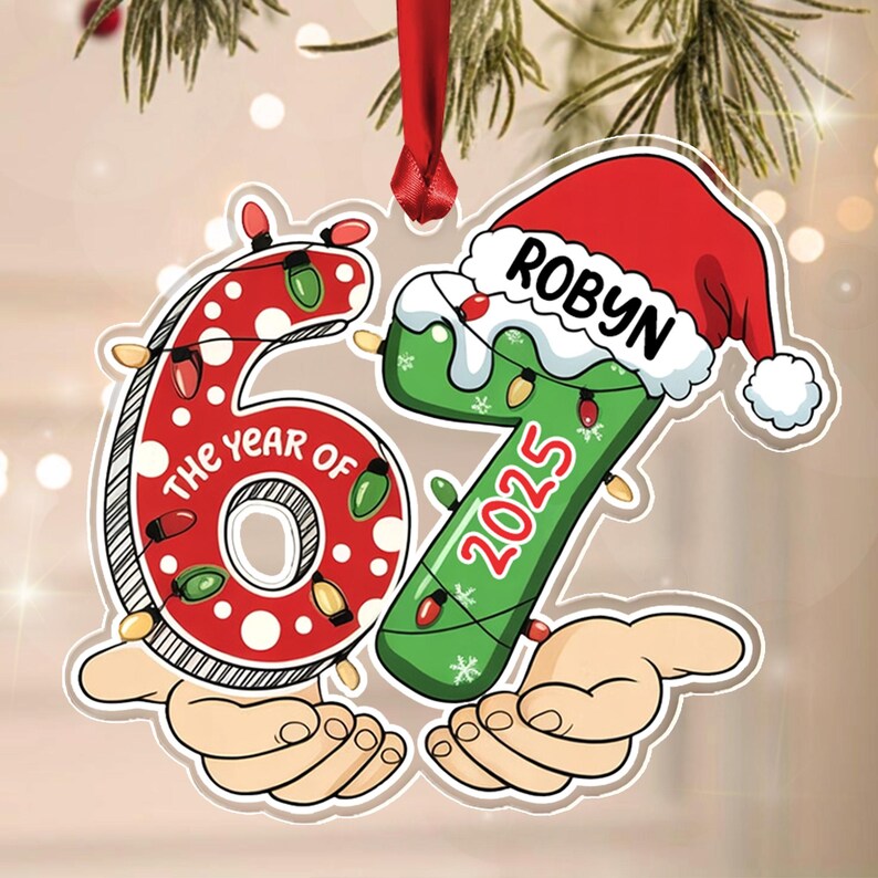A Christmas ornament shaped like the number 67, featuring a red polka-dot 6 and a green 7 wearing a Santa hat. The text reads THE YEAR OF and 2025, with the name ROBYN on the hat. It is decorated with colorful string lights.
