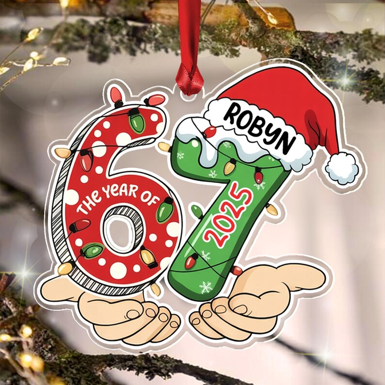 A flat, die-cut Christmas ornament shaped like the number 67. The red number 6 features white polka dots and string lights, with the text THE YEAR OF. The green number 7 wears a Santa hat with the name ROBYN and the year 2025.