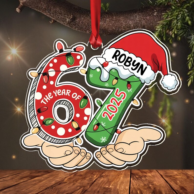 A flat, hanging Christmas ornament shaped like the number 67. The red number 6 features white polka dots and string lights, while the green number 7 wears a Santa hat with the name ROBYN and the year 2025 printed on it.