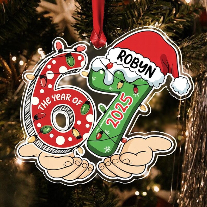 A Christmas tree ornament shaped like the number 67, featuring a red polka-dot 6 and a green 7 wearing a Santa hat. The text reads THE YEAR OF, ROBYN, and 2025. The design includes colorful string lights and two cartoon hands.