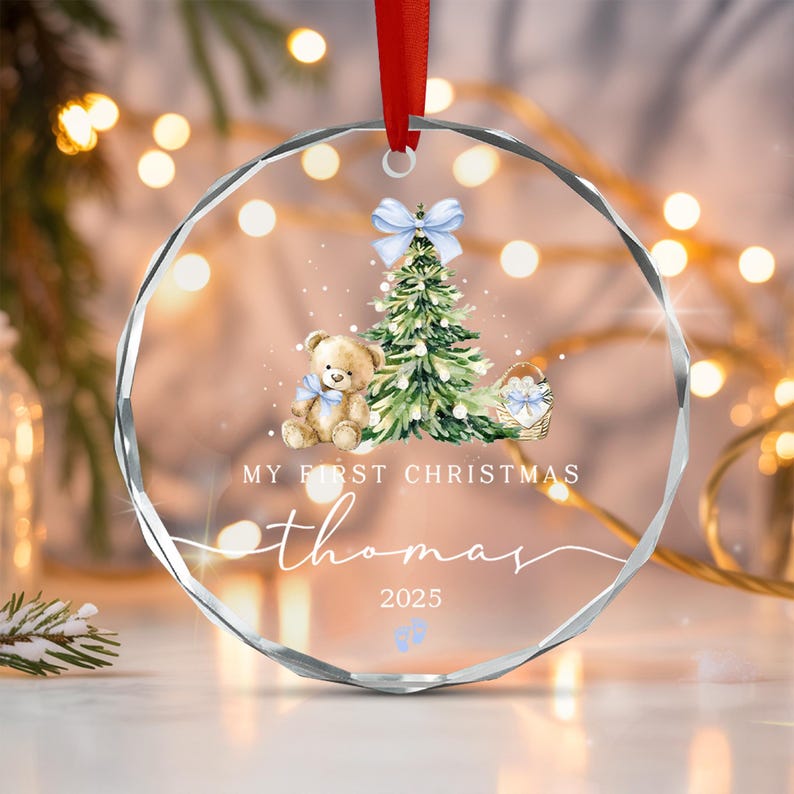 A circular clear acrylic Christmas ornament with a red ribbon hanger. The design features a teddy bear, a Christmas tree, a basket, and the text "MY FIRST CHRISTMAS", "thomas", "2025", and two small blue baby footprints.