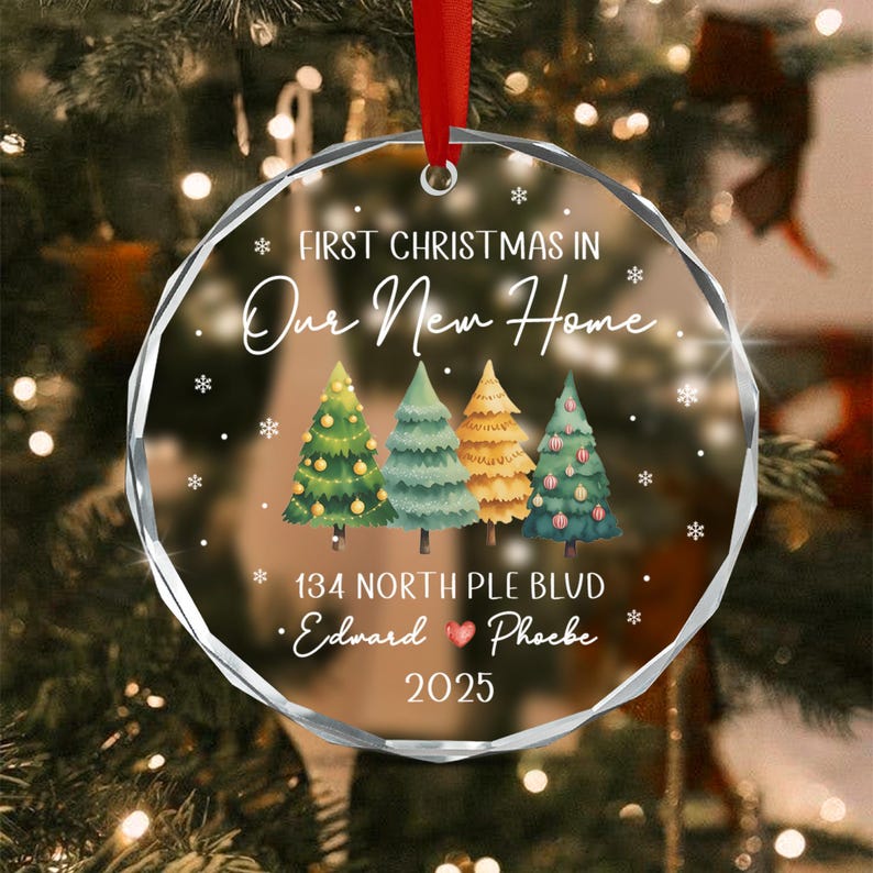 A round, clear acrylic Christmas ornament with a red ribbon hanger. It features the text "FIRST CHRISTMAS IN Our New Home", "134 NORTH PLE BLVD", "Edward & Phoebe", and "2025".