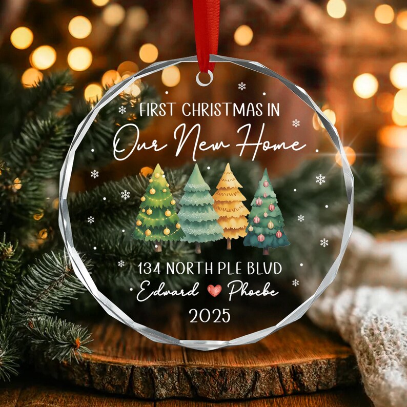 A round, clear acrylic Christmas ornament featuring the text First Christmas in Our New Home, 134 North Ple Blvd, Edward, a heart icon, Phoebe, and 2025.