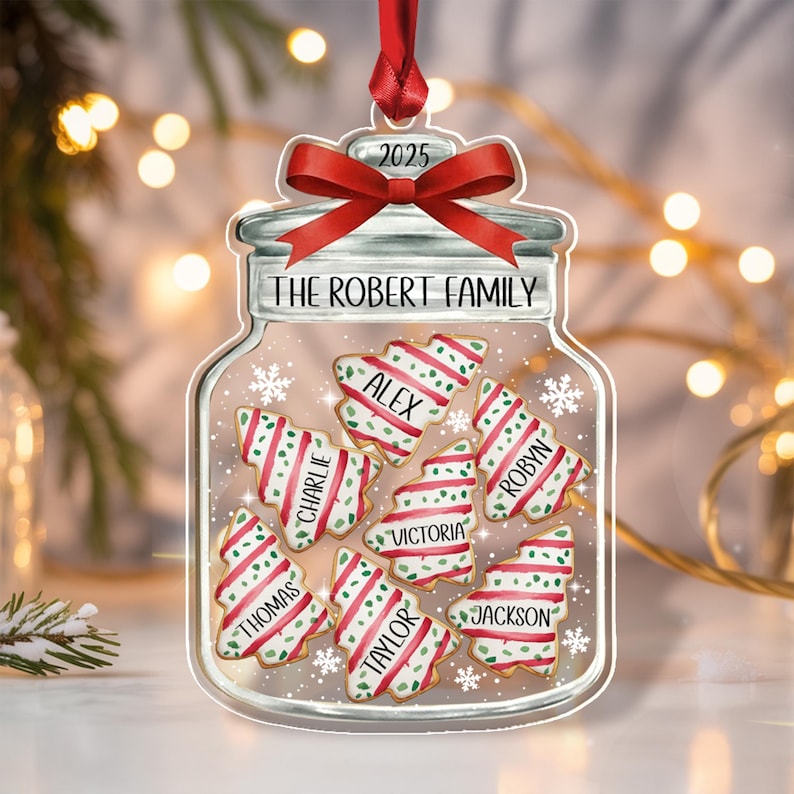 A clear, jar-shaped Christmas ornament featuring a red ribbon bow at the top and the text 2025. The jar is labeled THE ROBERT FAMILY and contains six tree-shaped cookies with red and green stripes, each personalized with names.