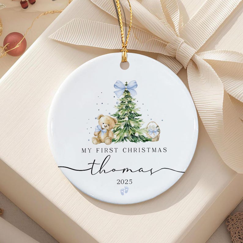 A round white ceramic ornament featuring a watercolor illustration of a teddy bear, a Christmas tree with a blue bow, and a small basket.