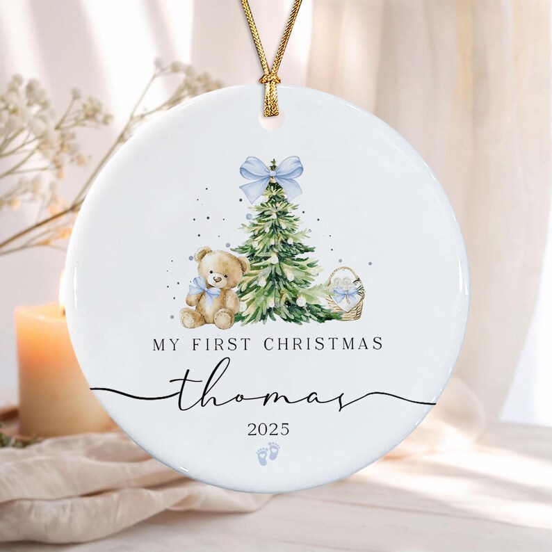 A round white ceramic ornament hanging from a gold cord. The design features a watercolor teddy bear, a green Christmas tree with a blue bow, and a small basket.