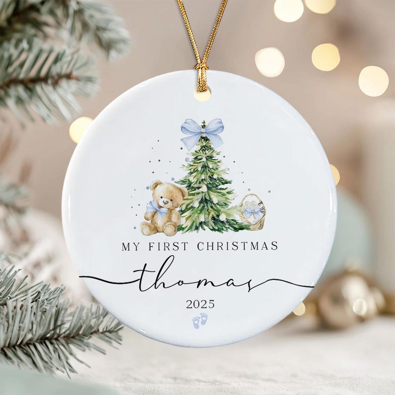A round white ceramic Christmas ornament hanging from a gold cord. The design features a watercolor teddy bear with a blue bow, a green Christmas tree with a blue bow topper, a basket, and the text My First Christmas Thomas 2025.