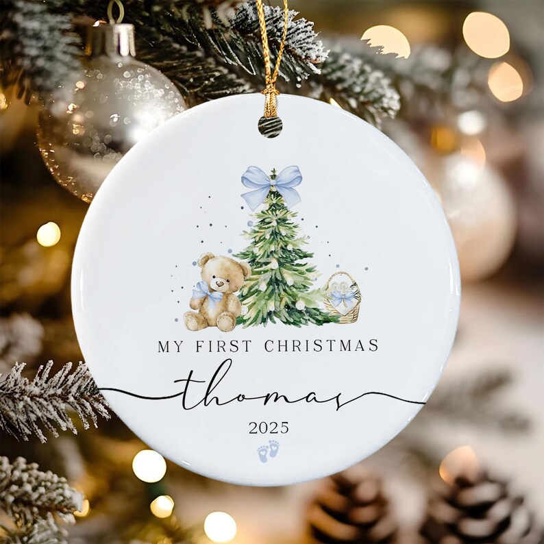 A round white ceramic ornament featuring a watercolor illustration of a green Christmas tree with a light blue bow, a brown teddy bear with a blue bow, and a basket.