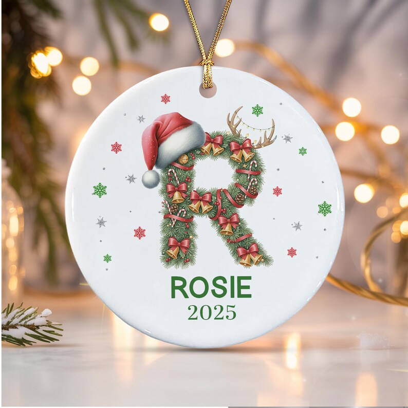 A round white ceramic ornament featuring a large letter R shaped like a pine wreath decorated with red bows, gold bells, and candy canes. A red Santa hat sits on top of the R. The name ROSIE and the year 2025 are printed in green.