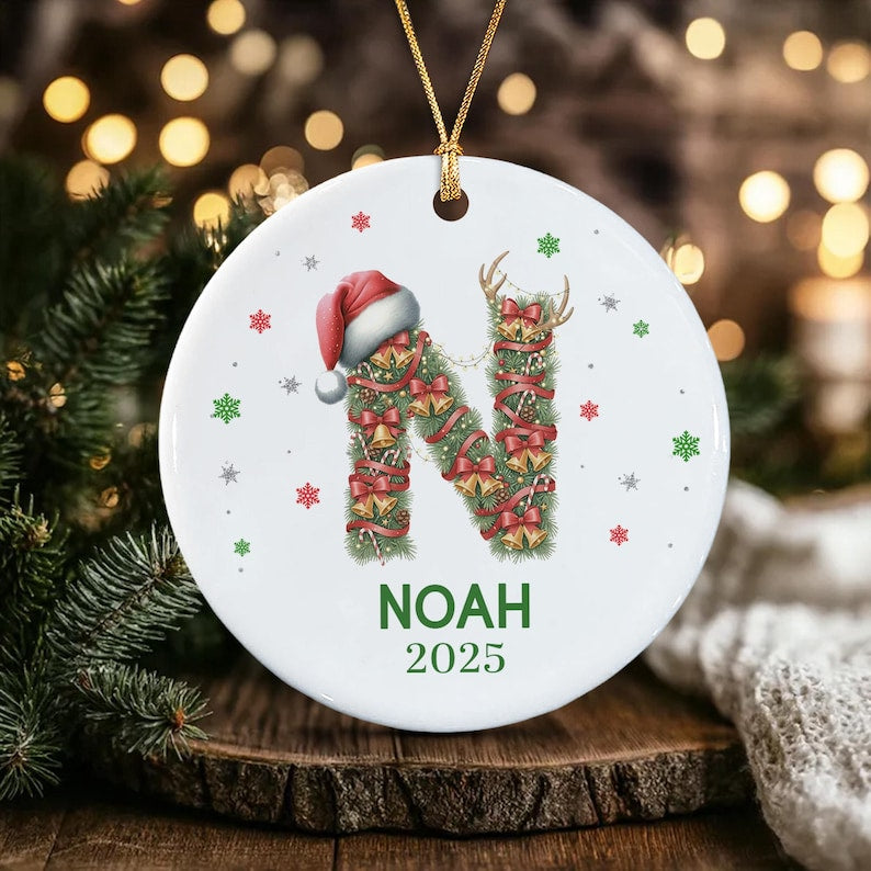 A round white ceramic ornament featuring a large letter N constructed from green pine branches, red ribbons, and gold bells. A red Santa hat sits atop the letter. The name NOAH and the year 2025 are printed below in green text.
