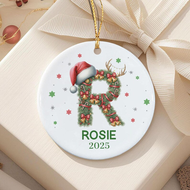 A round white ceramic Christmas ornament featuring a green pine wreath shaped like the letter R, decorated with small red bows, gold bells, and candy canes.