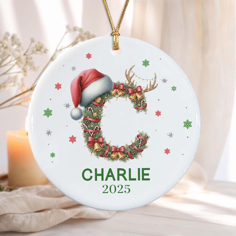 A round white ceramic ornament featuring a large letter C formed from a green pine wreath decorated with red bows, gold bells, and candy canes. A red Santa hat sits on top of the C, which has reindeer antlers.
