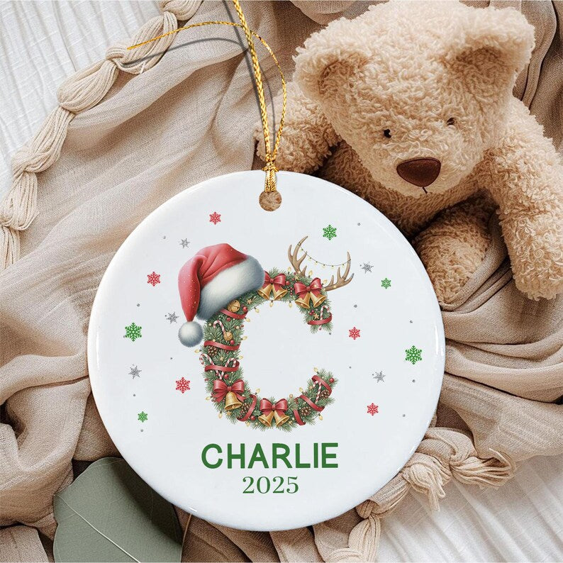 A round white ceramic ornament featuring a large letter C decorated with pine branches, red bows, gold bells, and a Santa hat.