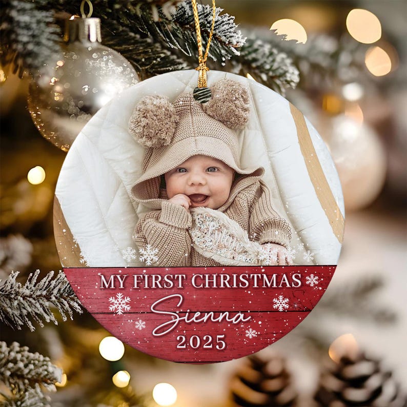 A round ceramic Christmas ornament featuring a photograph of a baby wearing a tan hooded sweater with pom-poms. The bottom section has a red wood-grain background with white snowflakes and the text MY FIRST CHRISTMAS Sienna 2025.