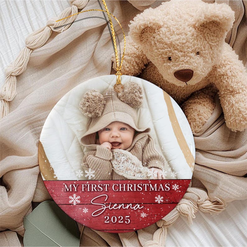 A round ceramic Christmas ornament featuring a photograph of a baby wearing a beige hooded sweater with pom-poms.