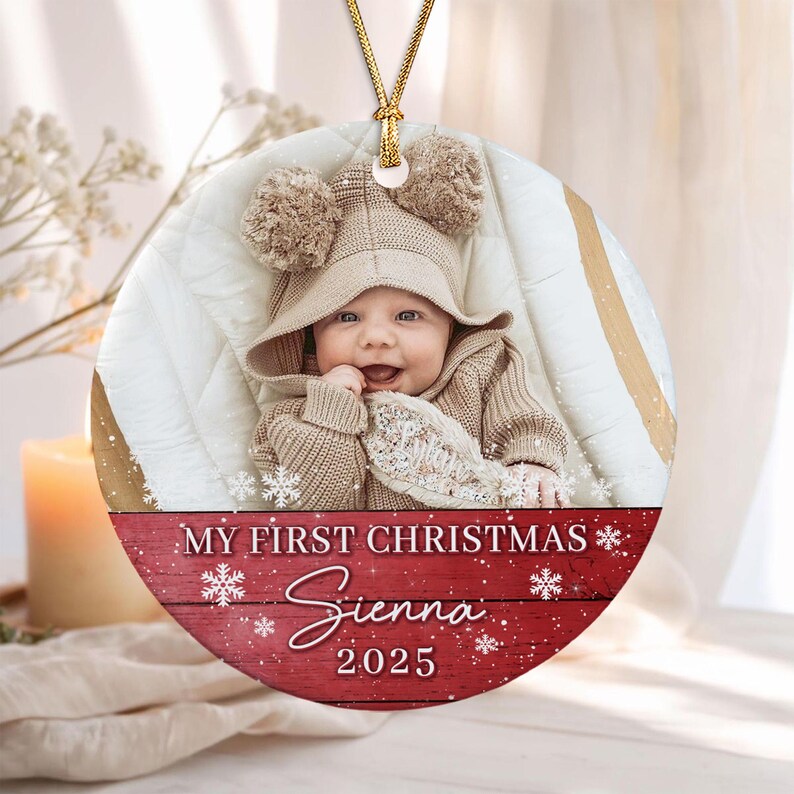 A round ceramic Christmas ornament featuring a photograph of a baby wearing a beige knit hat with two pom-poms. The bottom section has a red wood-grain background with white snowflakes and the text: MY FIRST CHRISTMAS Sienna 2025.