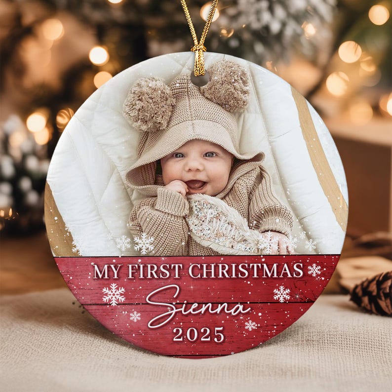 A round ceramic ornament featuring a photograph of a baby wearing a beige knit hat with pom-poms. The bottom section has a red wood-grain background with white snowflakes and the text: MY FIRST CHRISTMAS, Sienna, and 2025.