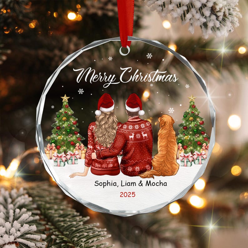 A circular glass Christmas ornament featuring a printed illustration of two people and a golden retriever dog sitting before two decorated Christmas trees.