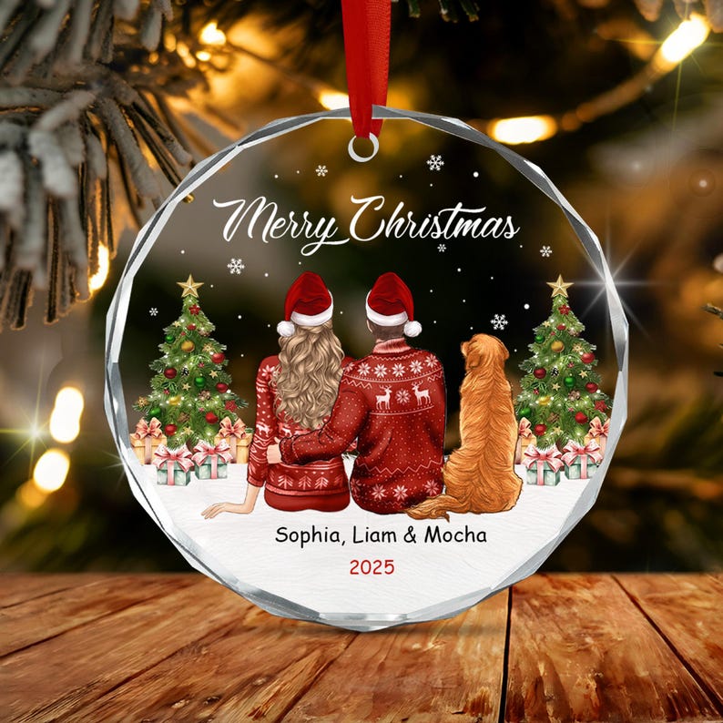A round, clear glass Christmas ornament featuring a printed illustration of two people and a golden dog sitting between two decorated Christmas trees. The text reads Merry Christmas, Sophia, Liam & Mocha, and the year 2025.