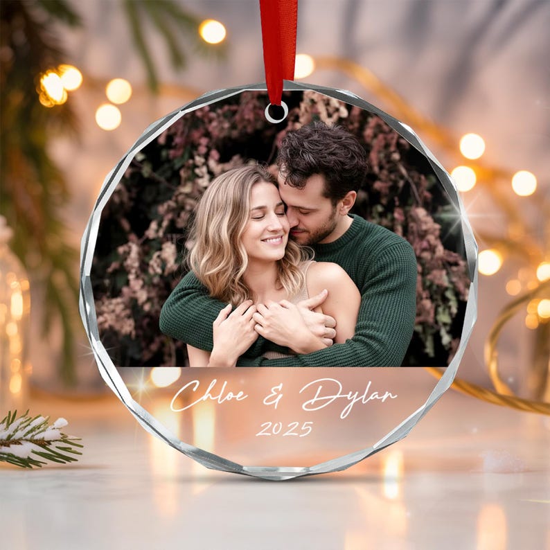 A circular glass ornament with faceted edges, featuring a printed photograph of a couple embracing. Below the image, the text "Chloe & Dylan" and "2025" is printed in white script. A red ribbon is attached to the top for hanging.