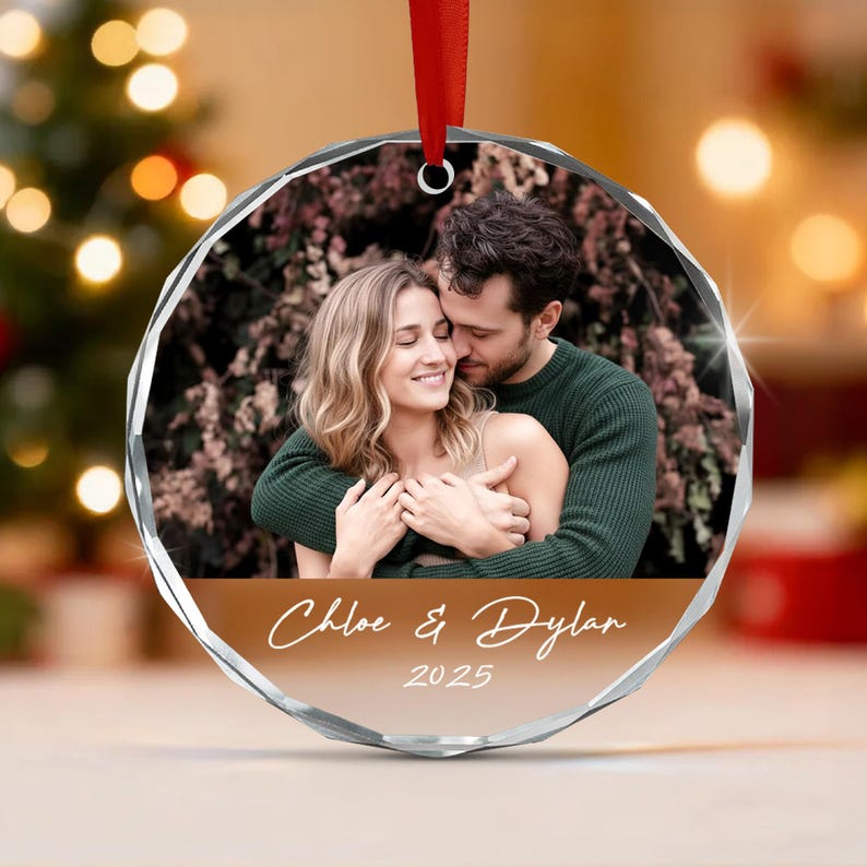 A circular faceted glass ornament featuring a photograph of a couple embracing. The bottom section has a solid brown background with the white text Chloe & Dylan and the year 2025. A red ribbon is attached to the top for hanging.