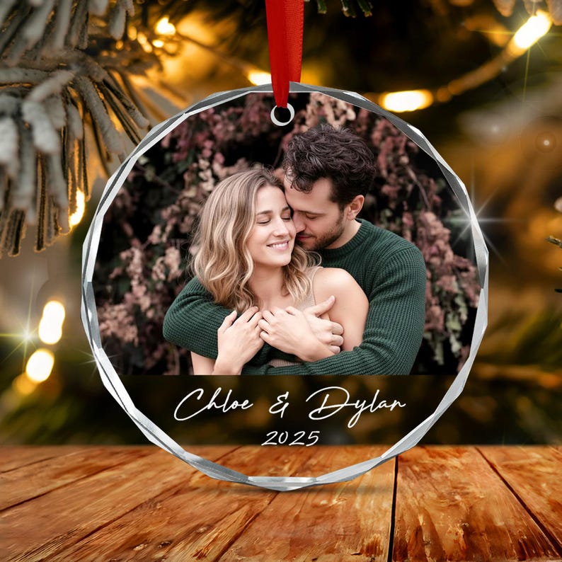 A circular, faceted glass Christmas ornament featuring a printed photograph of a couple embracing. The text "Chloe & Dylan" and "2025" is printed in white script below the image. A red ribbon is attached to the top for hanging.