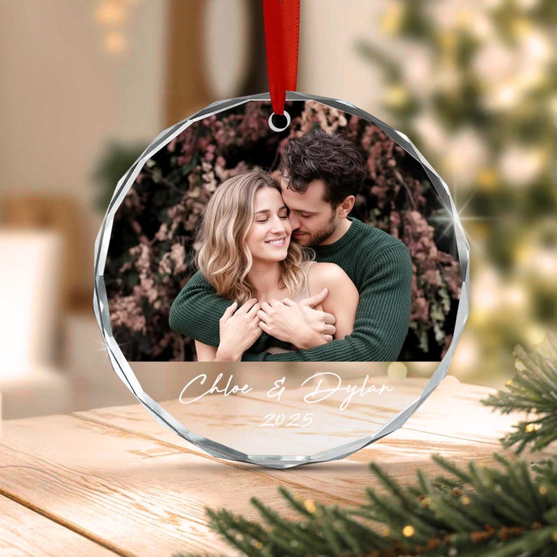 A circular glass ornament with faceted edges featuring a photograph of a couple embracing. The text "Chloe & Dylan" and "2025" is printed in white script on the lower portion. A red ribbon is attached to the top for hanging.