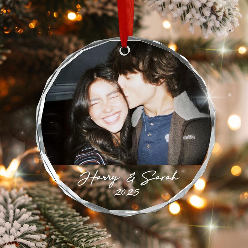 A circular glass ornament featuring a photograph of two people, with the text Harry & Sarah and the year 2025 printed in white script below the image. The ornament is suspended by a red ribbon and has a faceted, clear edge.