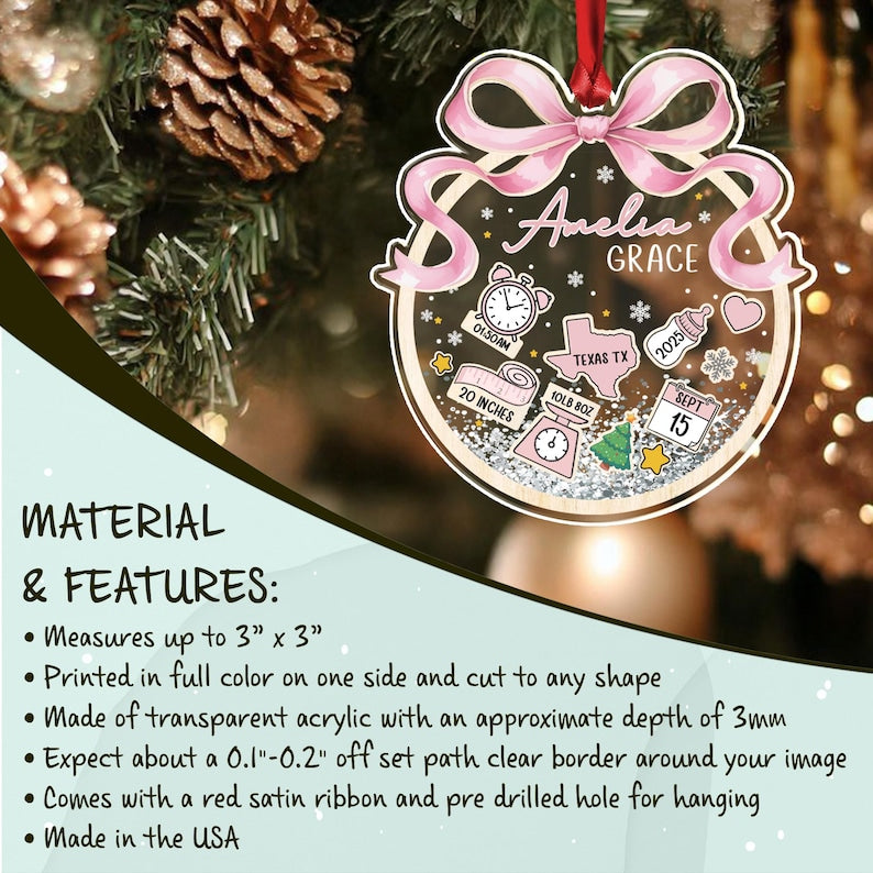 A personalized acrylic ornament with the name "Amelia Grace" in pink script. The ornament features baby-related icons, including a clock, measuring tape, and a baby bottle.