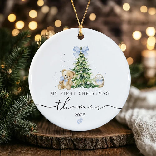 A round white ceramic Christmas ornament hanging by a gold cord. The design features a watercolor teddy bear, a decorated pine tree with a blue bow, and a small basket.