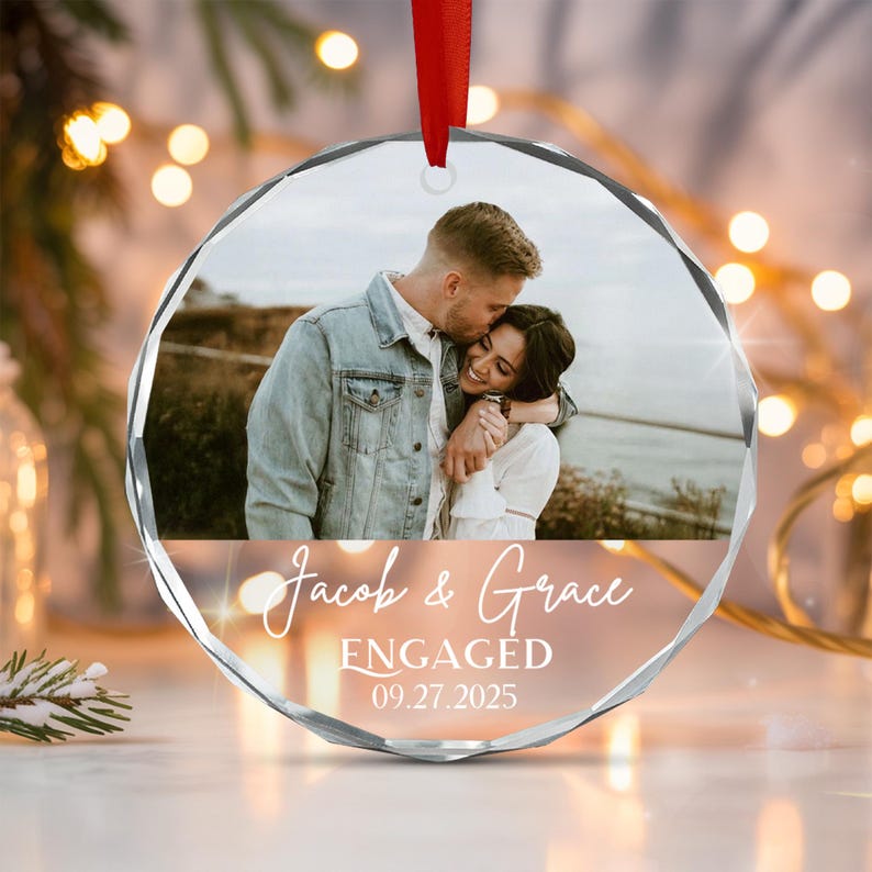 A circular faceted glass ornament featuring a photograph of a couple embracing outdoors. The text Jacob & Grace ENGAGED 09.27.2025 is printed in white below the photo. A red ribbon is attached to the top for hanging purposes.